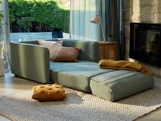 A light green sofa with a tan and white pillow, next to a woven basket and a modern lamp, set in a cozy room with a fireplace.