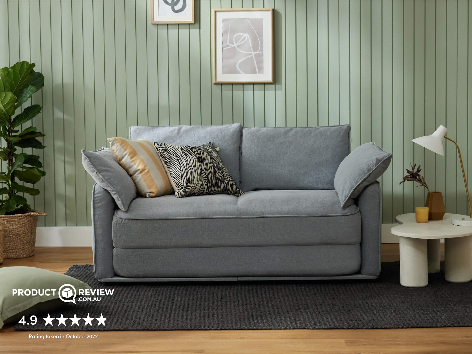 A light grey sofa with varying cushions, including a striped golden one, on a textured dark grey rug. A green wall and contemporary lamp complete the scene.