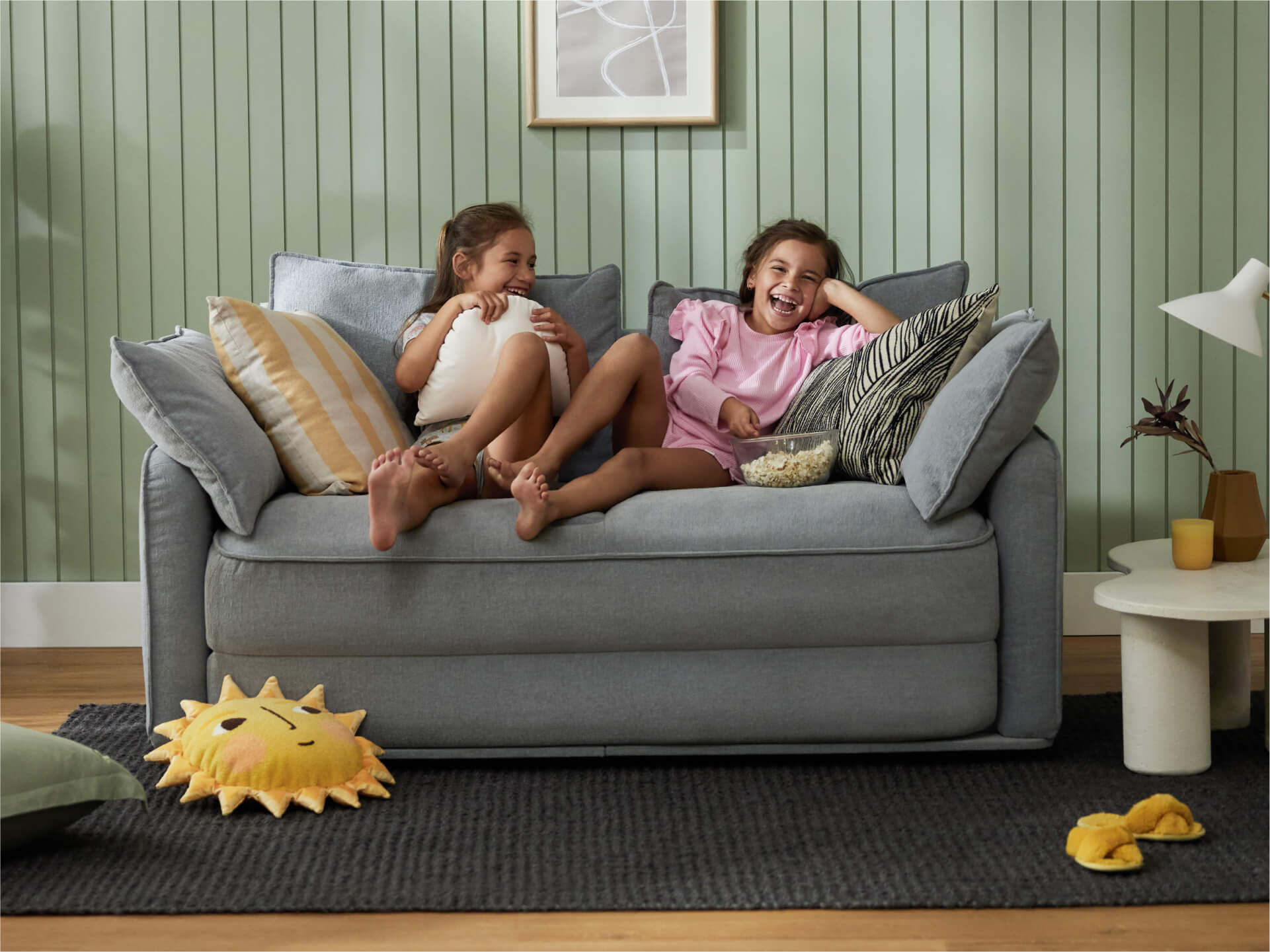 Two girls sit on a light grey sofa with blue and striped cushions, laughing. A yellow sun pillow rests on a dark rug nearby.