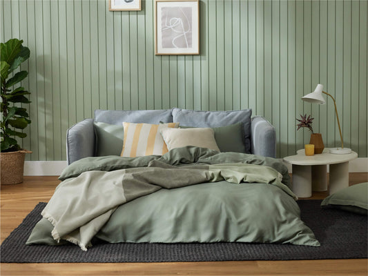 Cozy room featuring a light grey sofa with green and beige pillows, a green wall, a cream rug, and a white lamp on a round table.