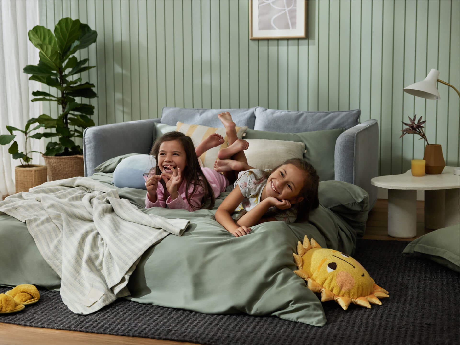 Two girls lie on a light green bedspread, laughing. A light blue sofa and a potted plant are behind them, with a sun-shaped pillow nearby.