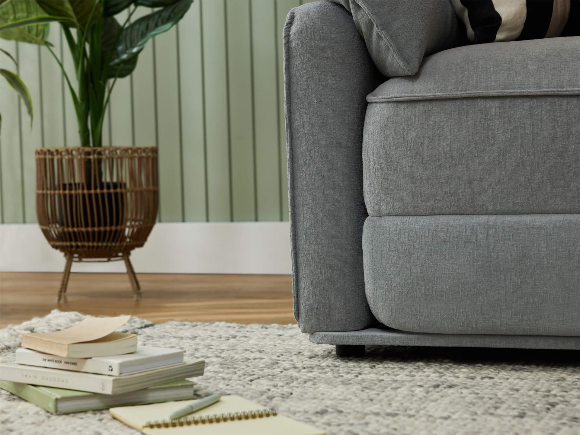 A light grey sofa with textured fabric sits beside a woven, dark brown plant basket. A beige and green patterned rug is beneath a stack of books.