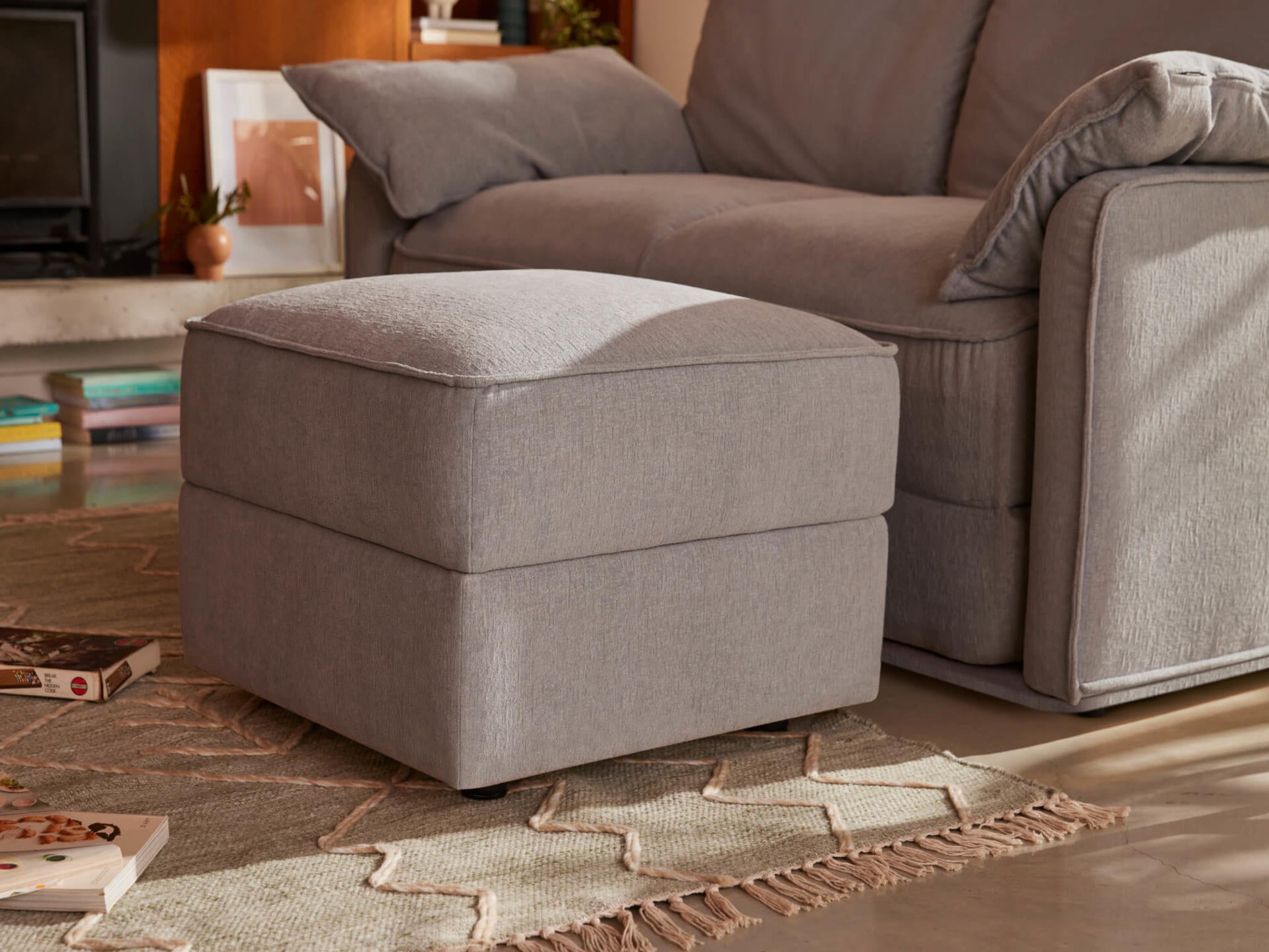 A light grey upholstered ottoman sits on a beige textured rug, next to a matching grey sofa with a fluffy throw pillow.