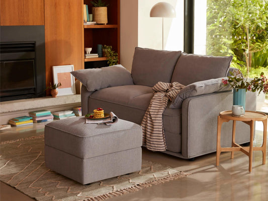 A light grey sofa with striped throw blanket, beside a dark grey ottoman topped with snacks. A wooden side table holds a vase with flowers.