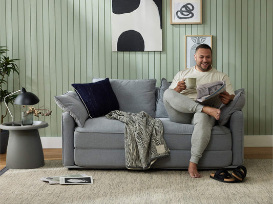 A man sits on a light grey sofa with a dark blue pillow, holding a green mug. The room features mint green walls and a dark lamp.