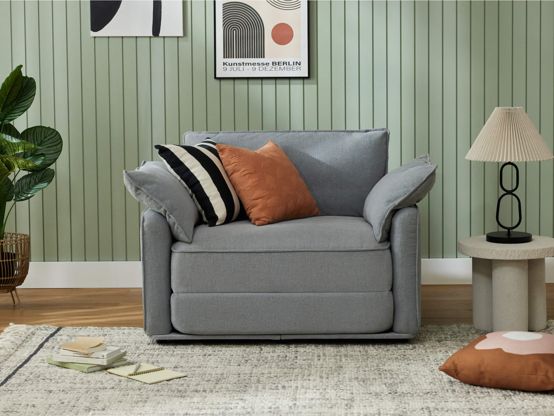 A light grey sofa with colorful cushions, a green paneled wall, a beige lamp, and a textured rug with a few books scattered nearby.