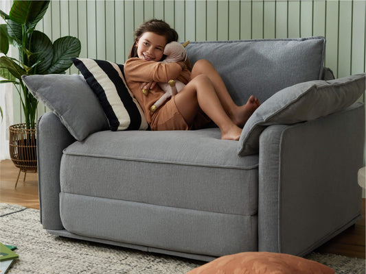 A young girl in a brown outfit smiles while hugging a stuffed animal on a light grey sofa, surrounded by green plants.