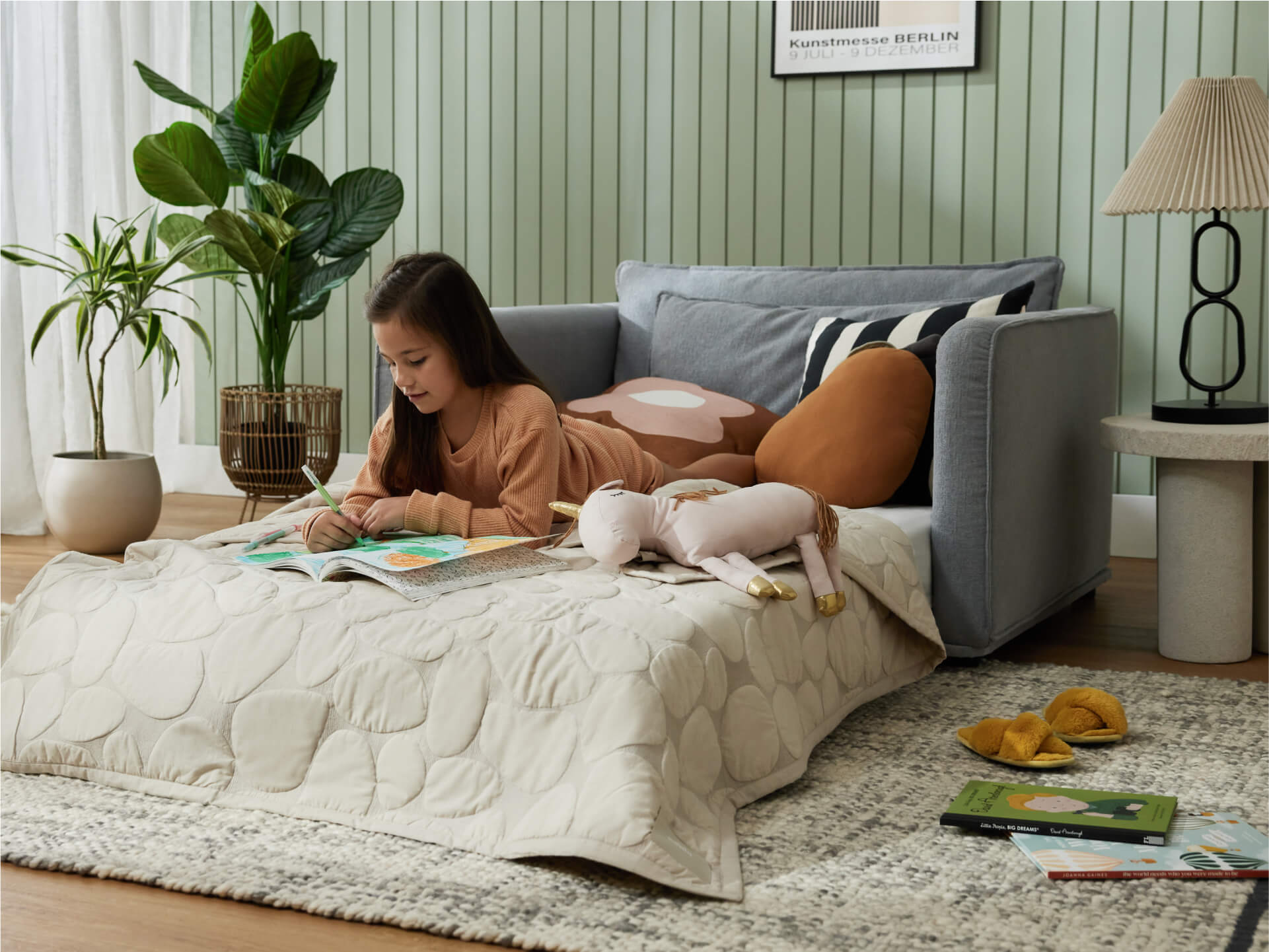 A girl in a beige sweater lays on a light gray sofa, coloring on a beige quilt. Nearby, plants, a brown pillow, and a lamp are visible.