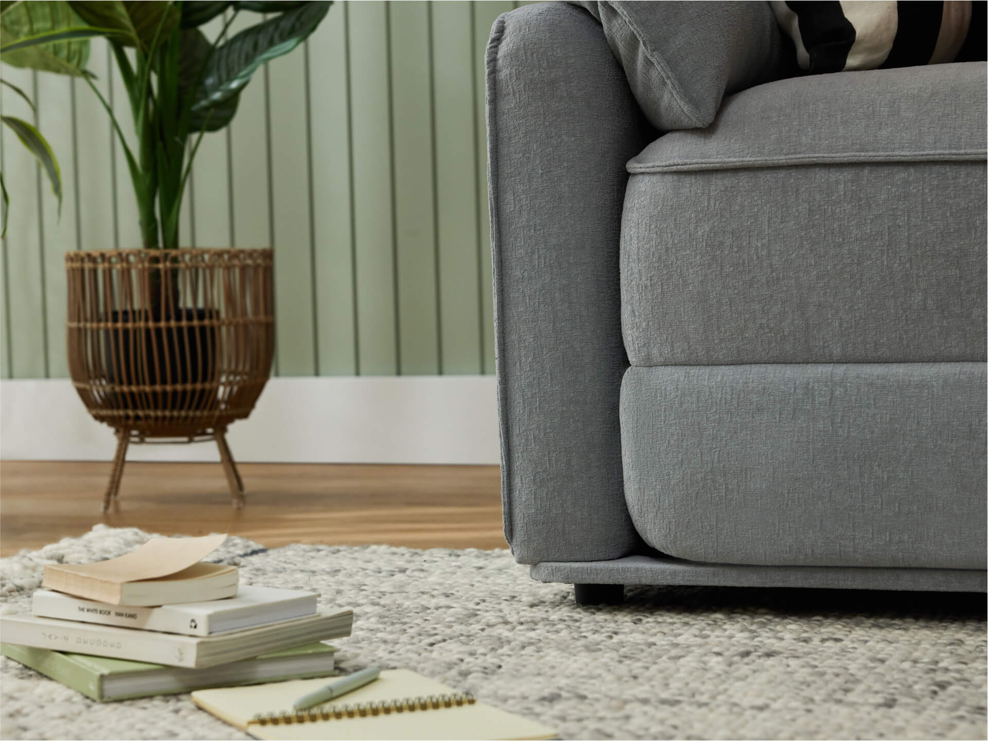 A light grey sofa sits beside a woven brown planter with green leaves, on a textured beige rug with stacked books and a notebook nearby.