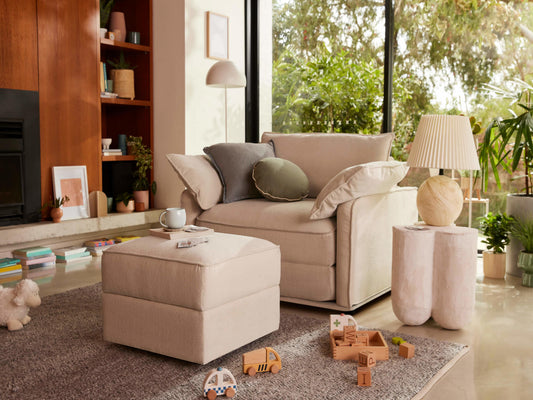 A light beige sofa with grey and green pillows sits beside a textured cream ottoman. Nearby, a white lamp, toys, and plants add color.