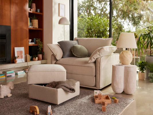 A beige sofa with green and grey cushions, a light gray ottoman, and a cream lamp on a white table, surrounded by plants and toys.