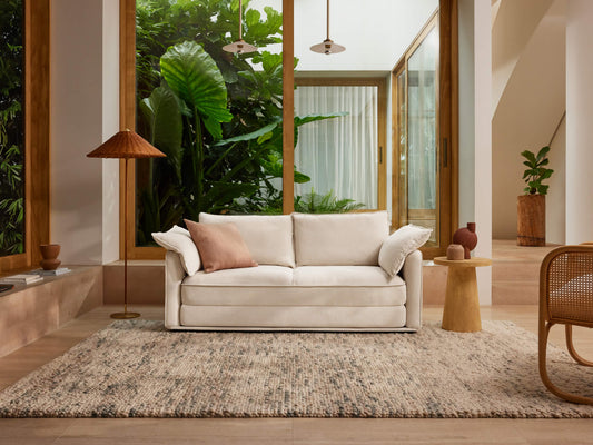 Bright living room featuring a white sofa with light pink cushion, tan rug, wooden accents, and lush green plants visible through large windows.