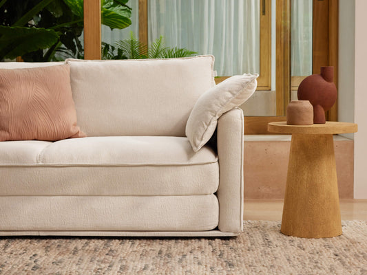 A light beige sofa with a textured cushion sits beside a round, light wood table. Two terracotta vases are on the table against a green backdrop.