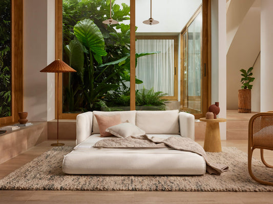 A white sofa with soft pillows rests on a beige rug in a bright, plant-filled room with large glass windows and wooden accents.