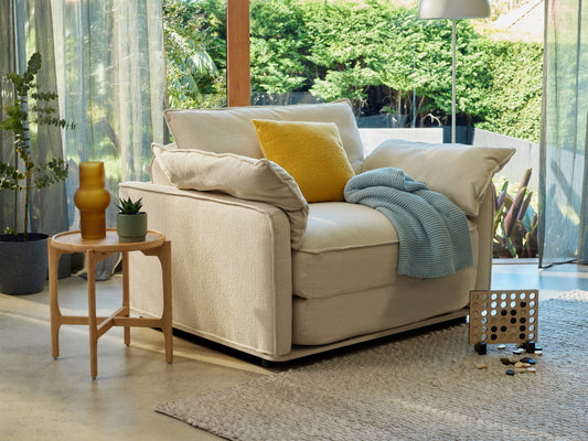 A light beige armchair with oversized cushions holds a yellow pillow and a blue throw. A wooden side table features a yellow vase and a small plant.