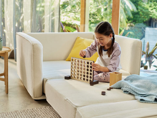 A young girl with braided hair plays a game on a cream-colored sofa. A light yellow pillow and a blue knitted blanket are nearby.