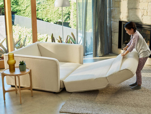 A light beige sofa, with a child in grey pajamas with pink polka dots, adjusting a cushion in a bright, modern living room.