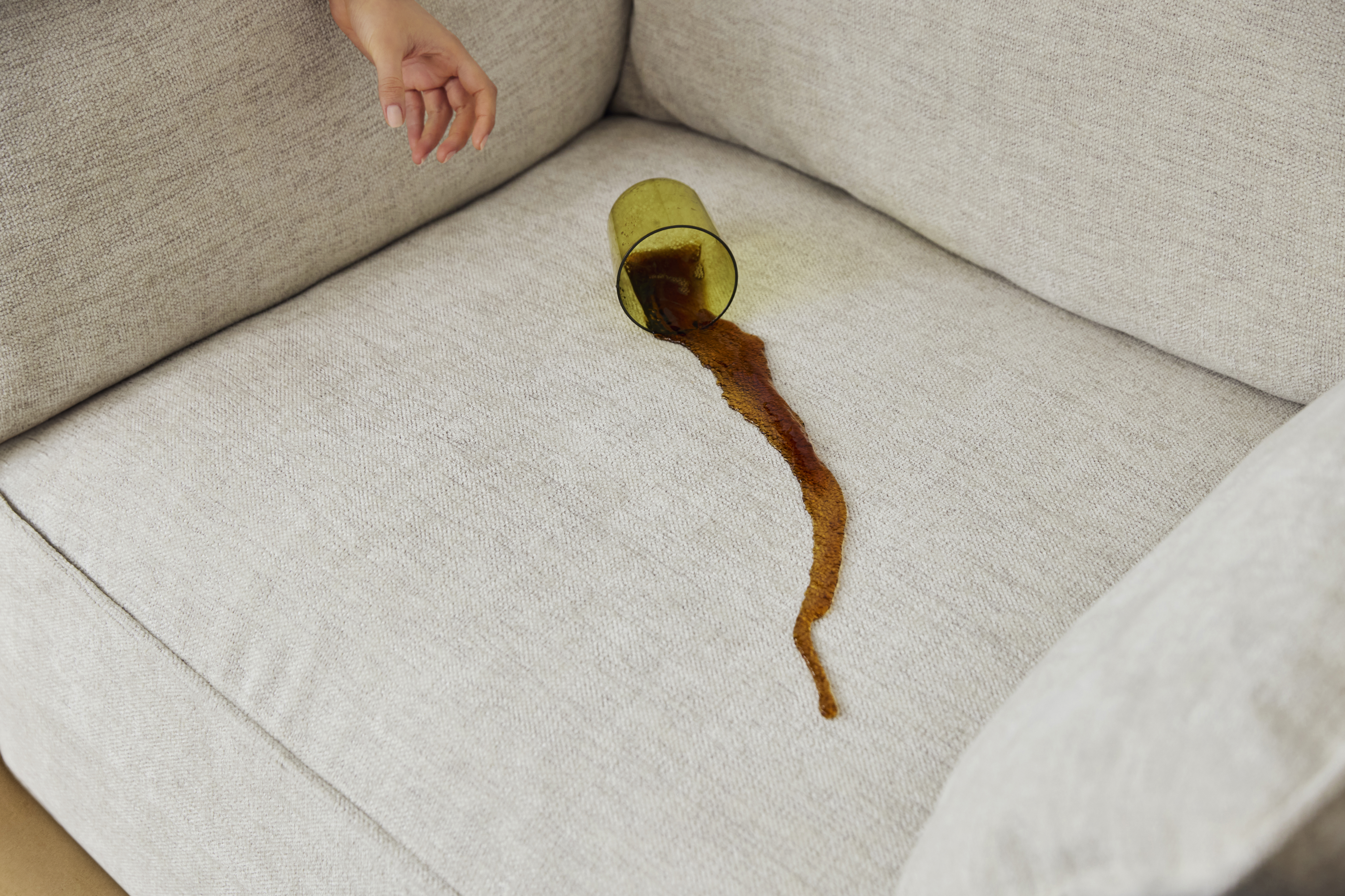 Glass of juice spilled on a beige sofa with a hand reaching towards it.