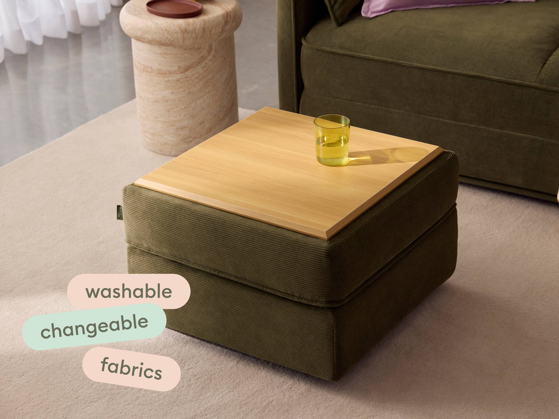 Modern ottoman with olive green washable fabrics and a light oak wood top. Features include a versatile, changeable design. Set in a contemporary living room.