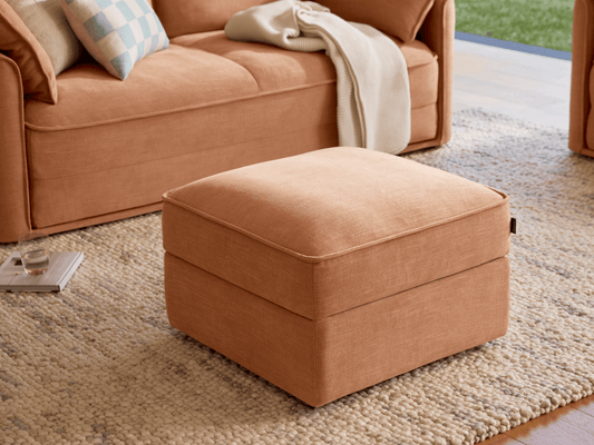 Luxurious ottoman in soft terracotta fabric, featuring a plush cushion and clean lines. Placed on a textured rug in an elegant, modern living space.