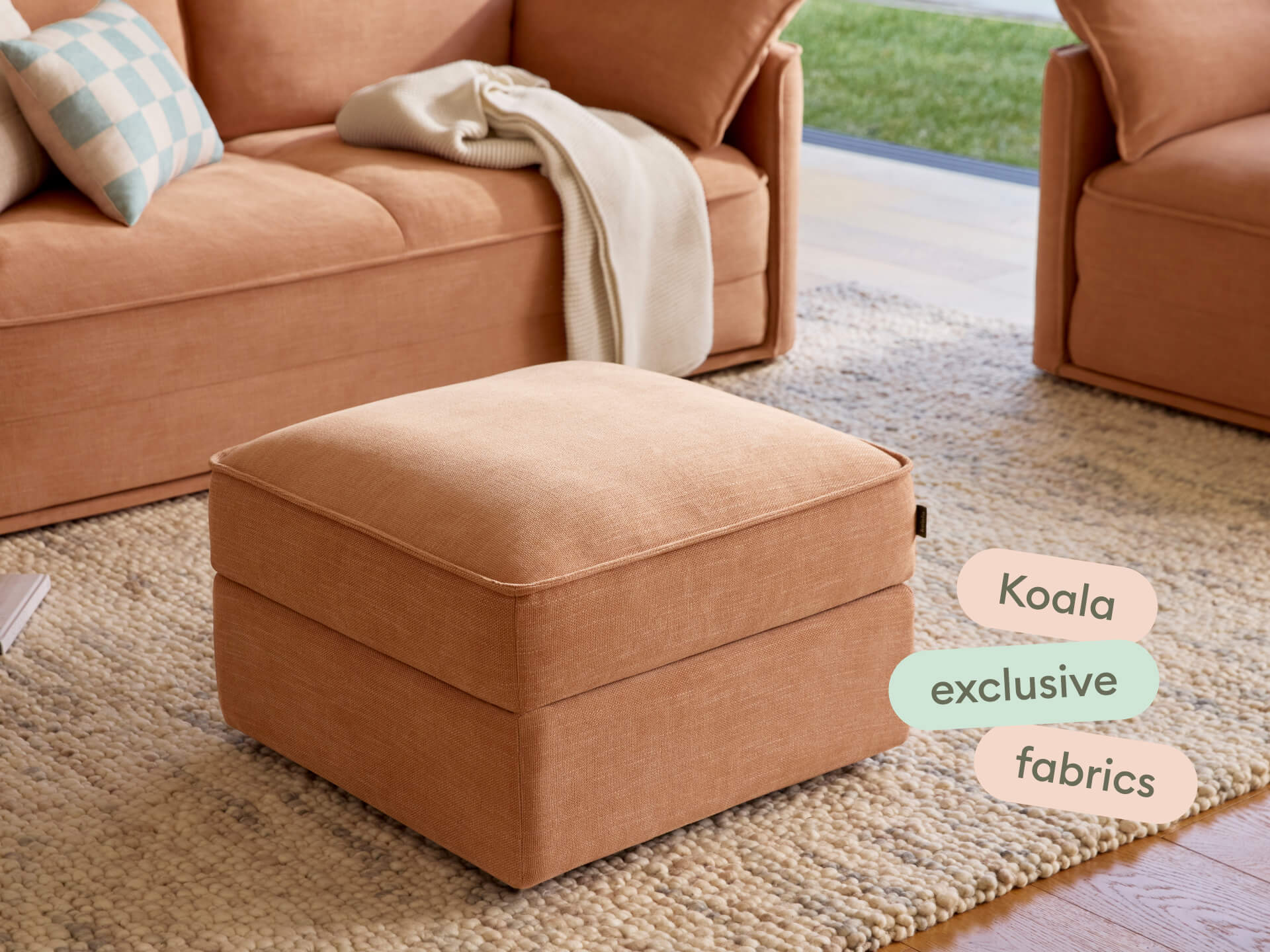 Luxurious ottoman in rich terracotta fabric, featuring clean lines and plush cushioning. Set on a textured neutral rug, complementing modern decor.