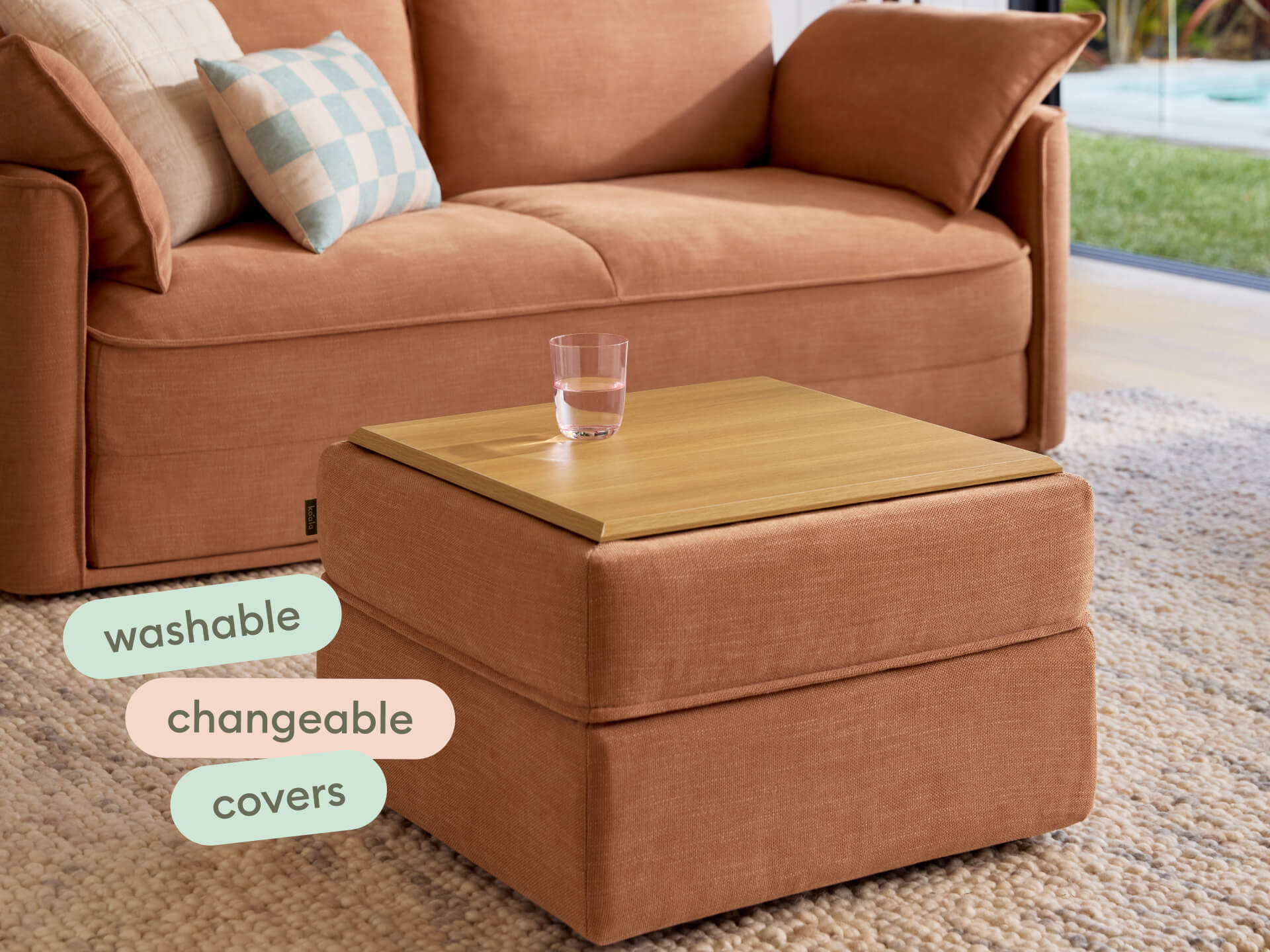 Elegant ottoman with terracotta fabric and washable, changeable covers; features a blonde wood top. Set in a modern living room with natural light.