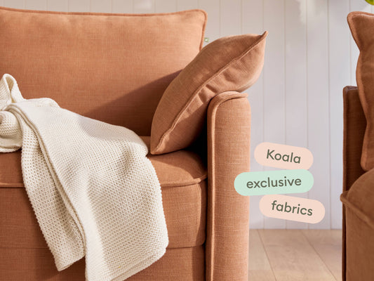 Rust-toned linen sofa with plush cushions, textured ivory throw draped over armrest. Set against light wooden floor and vertical white panelling.