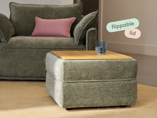 Square grey ottoman with a light wood flippable lid. Upholstered in textured fabric. Paired with a matching sofa in an elegant living room setting.