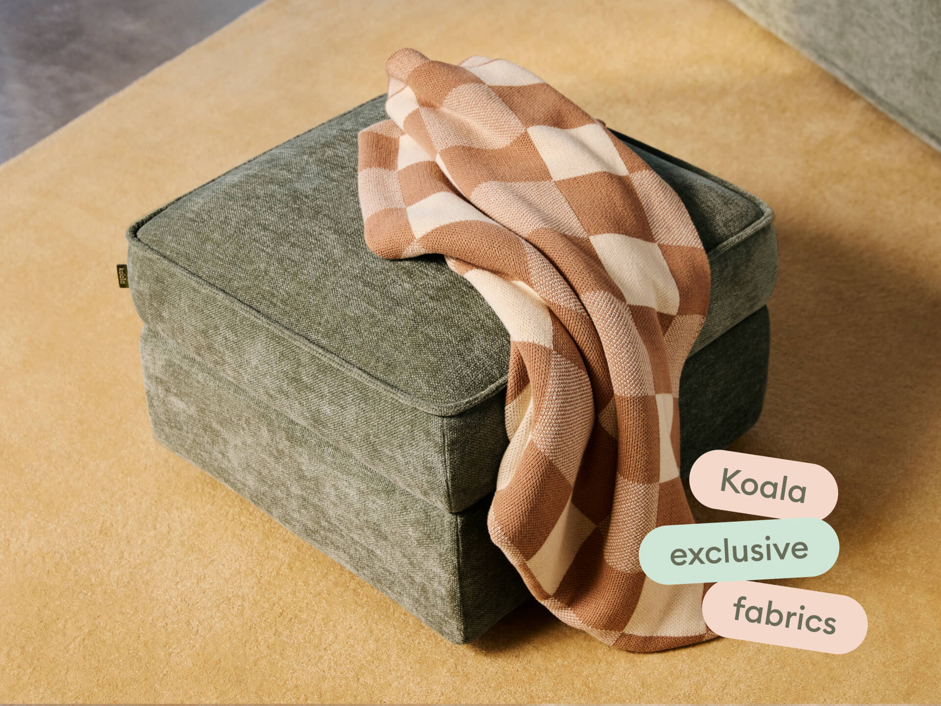 Grey cushioned ottoman with textured fabric, featuring clean lines and a soft finish, set on a beige rug. Highlighted by a draped checkered throw.