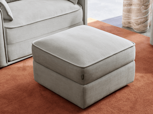 Elegant ottoman in light grey fabric with sleek piping detail, featuring a plush cushion top. Positioned on a rich terracotta rug in a sunlit room.