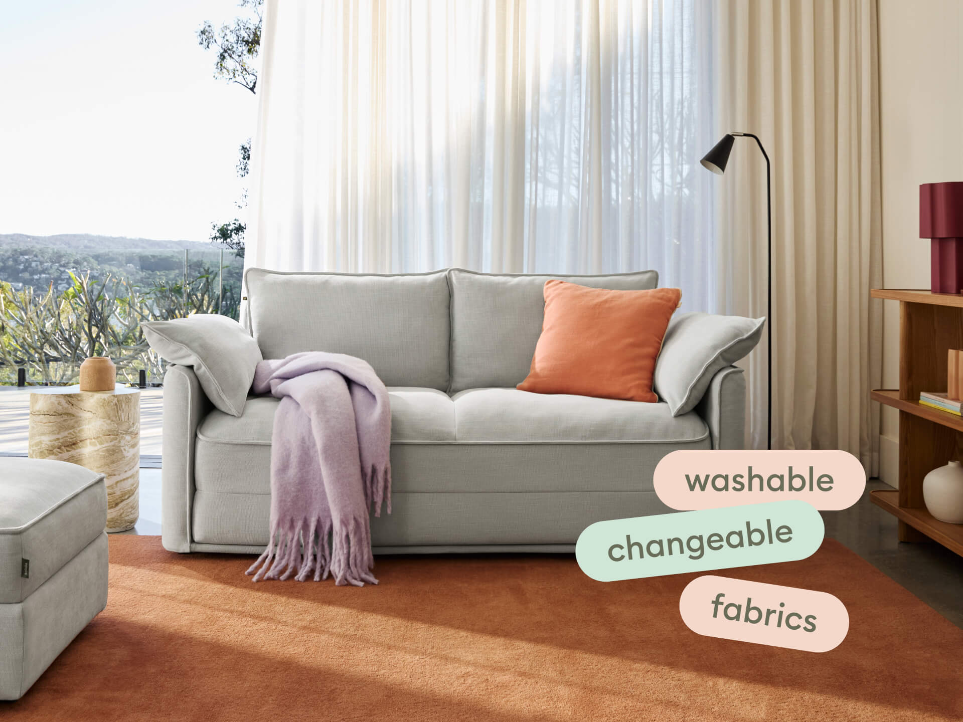 Luxurious grey fabric sofa with plush cushions, washable and changeable covers. Set against sheer curtains and a scenic landscape, exuding modern elegance.
