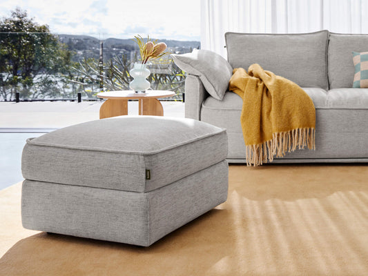 Premium grey fabric ottoman with a sleek upholstered design sits on a light brown carpet. It complements a plush sofa adorned with a mustard throw in a modern sunlit living space, with a glass table and scenic views.