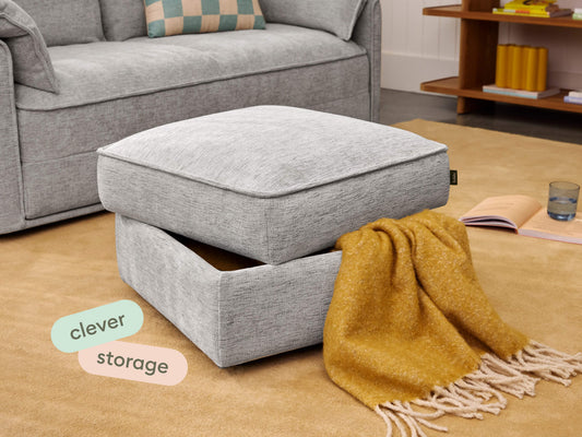 Modern grey ottoman with soft fabric upholstery, featuring hidden storage below a plush cushion. Set in a stylish living room on a light brown rug.