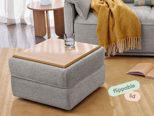 Modern grey fabric ottoman with a light wood finish flippable lid, set in a cosy living room with a plush sofa, mustard throw, and warm-toned rug.