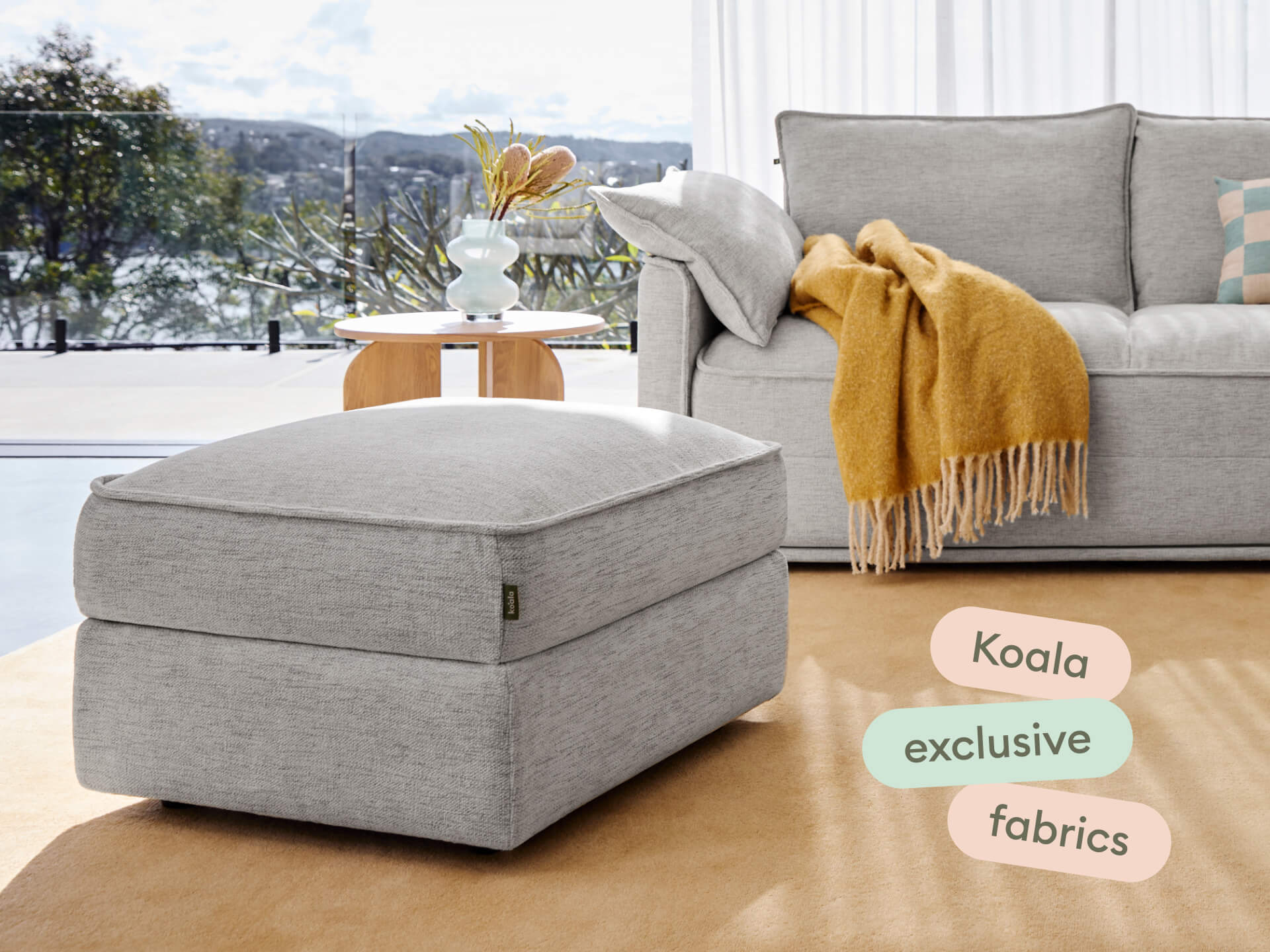 Elegant grey fabric ottoman with soft upholstery, smooth finish, and clean lines in a sunlit living room, complementing a plush sofa and wooden accents.