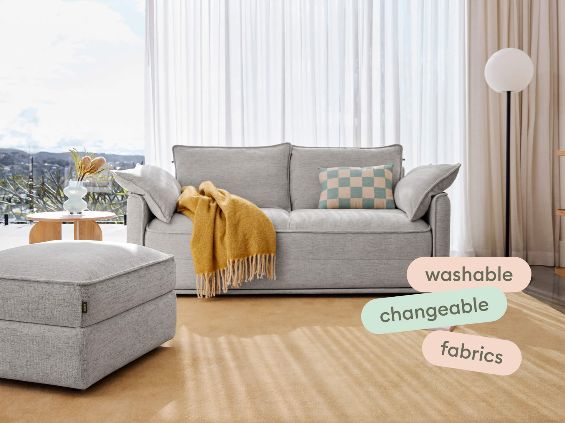 Luxurious grey fabric sofa with washable, changeable covers, paired with a matching ottoman. Set in a sunlit living room with sheer curtains and neutral decor.