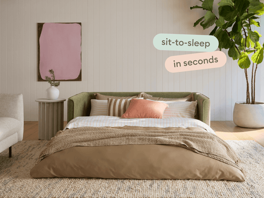 Luxurious green fabric sofa bed with sleek armrests, complemented by a tan duvet and striped pillows. Set in an elegant, light-filled room with a modern aesthetic.