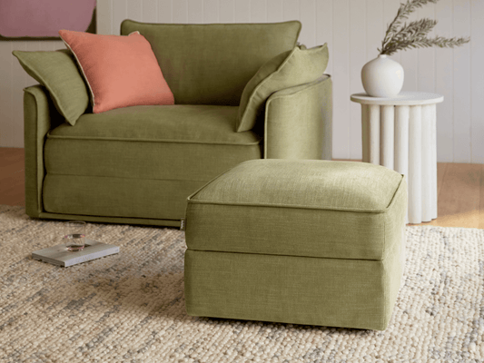 Luxurious olive green fabric armchair with matching ottoman, featuring plush cushions. Set on a textured woven rug in a sophisticated living space.