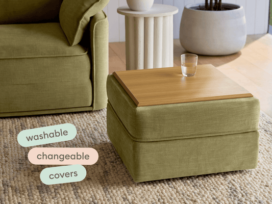 Olive green ottoman with a smooth timber top, featuring washable, interchangeable covers. Set on a textured rug beside a stylish armchair and ceramic pot.