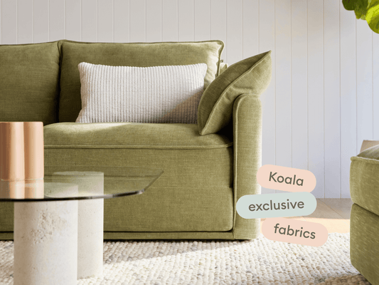 Luxurious olive-green fabric sofa with clean lines and plush cushioning, set on a woven rug. Features a glass coffee table in a serene, contemporary living room.