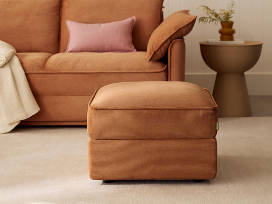Rust-coloured corduroy ottoman with plush cushioning, square shape, and modern design. Situated on a beige carpet beside a contemporary sofa and round side table.