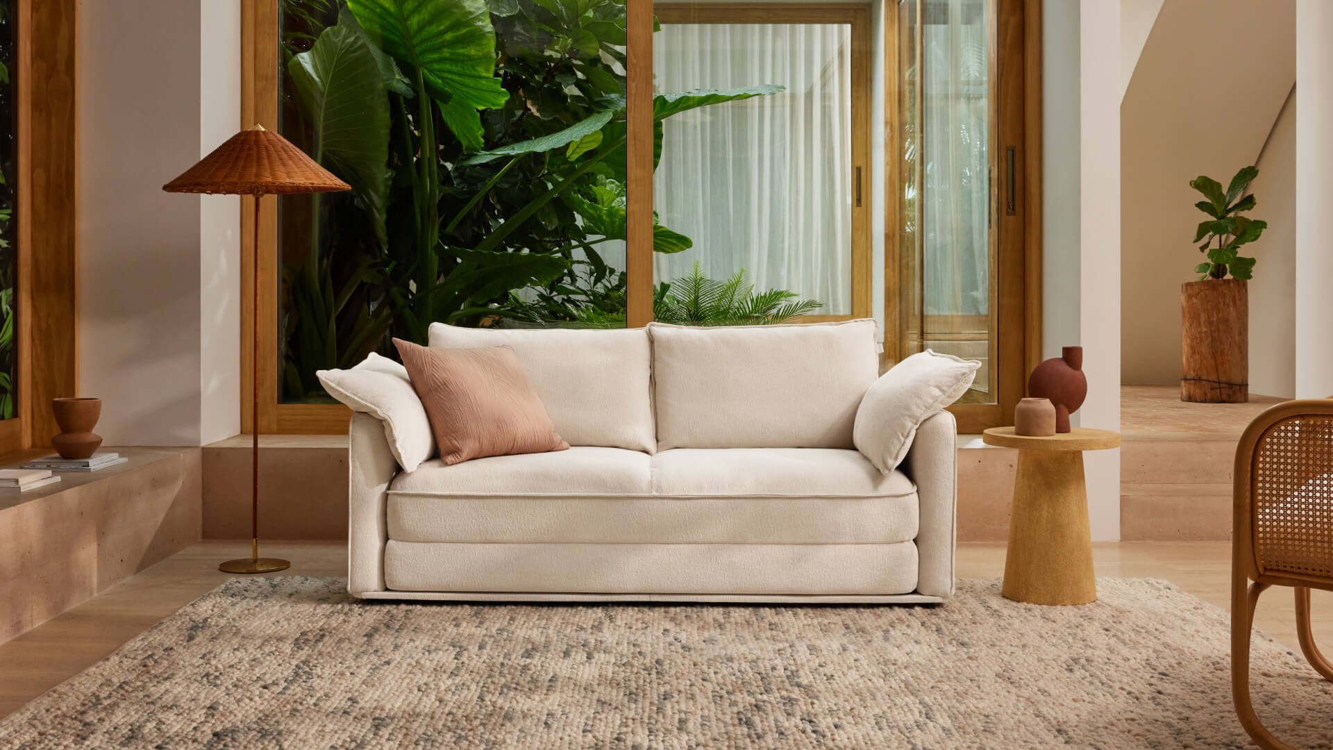 A light beige sofa with two cushions sits in a bright room surrounded by green plants, with a rattan lamp and a wooden side table nearby.