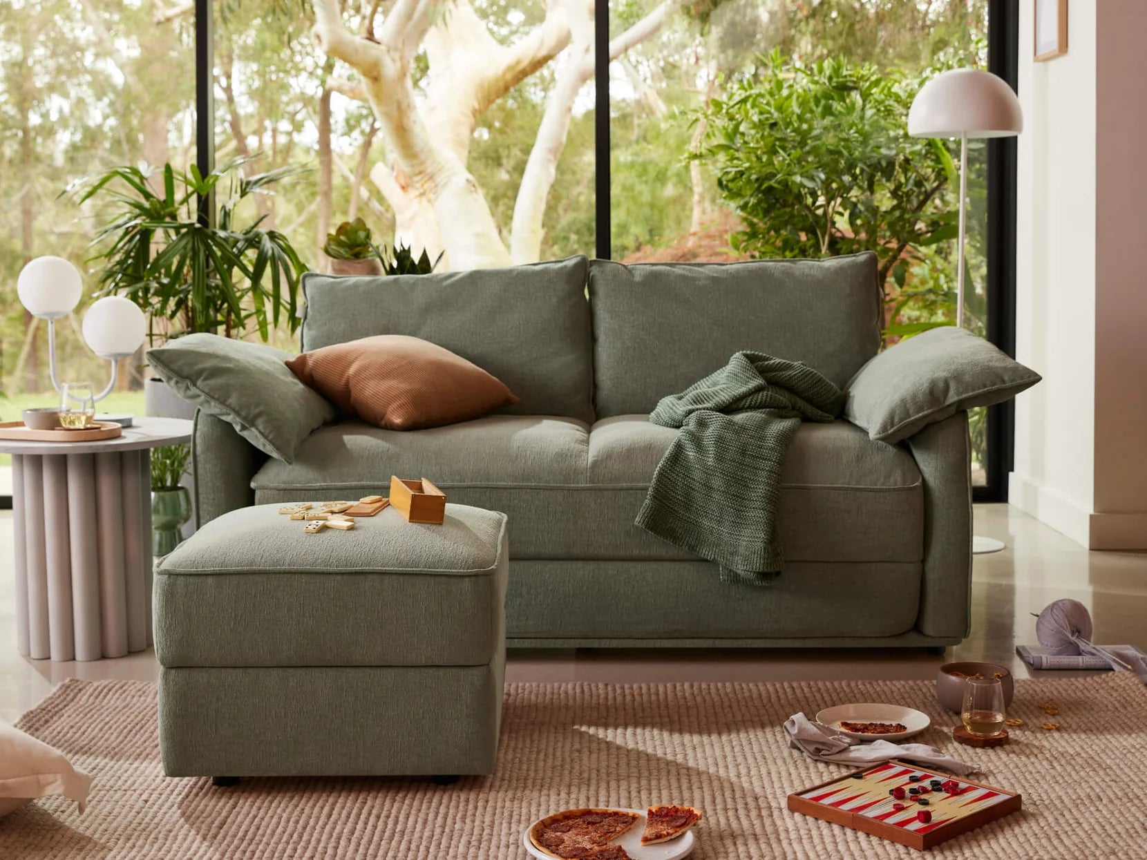 A light green sofa with two cushions sits beside a matching ottoman. A brown pillow and teal throw drape over the sofa, with snacks on a white rug.