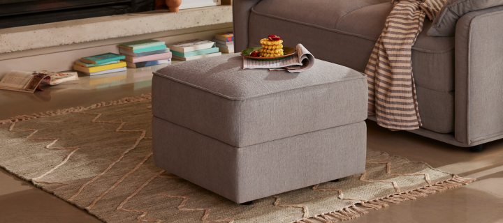 A light grey ottoman sits on a beige, patterned rug in a cozy living room, with a stack of books and a striped throw nearby.