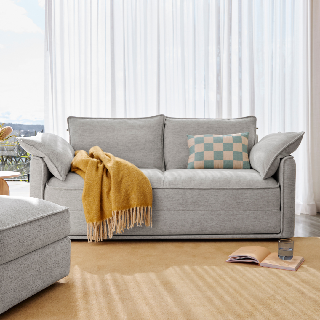 Modern grey fabric sofa with plush cushions, accented by a mustard throw and checkered pillow. Set on a beige carpet near sheer white curtains.