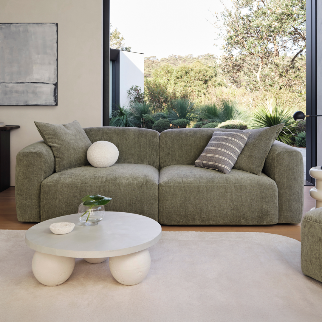 Olive green fabric sofa with plush cushions sits in a modern living room, featuring soft curves and a low profile. Positioned on a light hardwood floor.