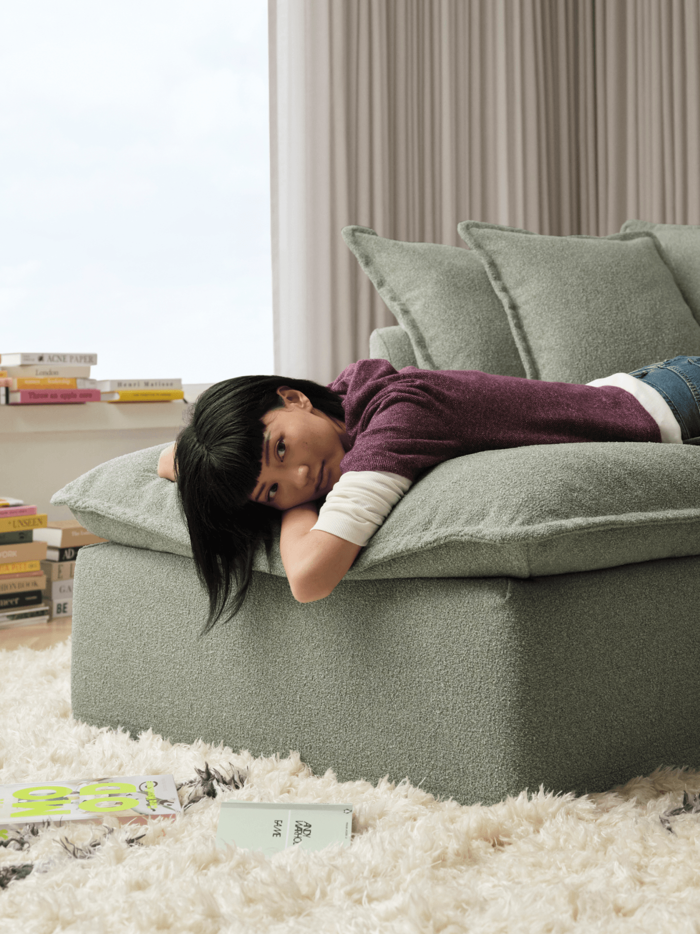Person lying on a green sofa in a living room with books and a window in the background