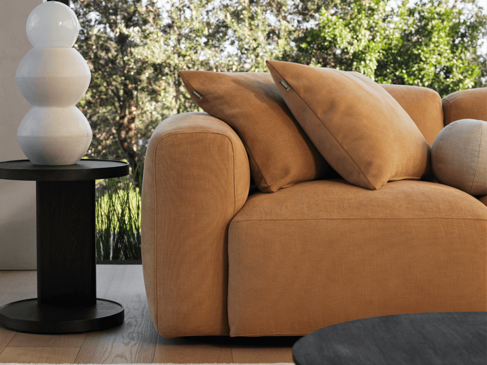 Luxurious tan linen sofa with plush cushions, set against a backdrop of lush greenery. Paired with a sleek black side table and a modern sculptural lamp.
