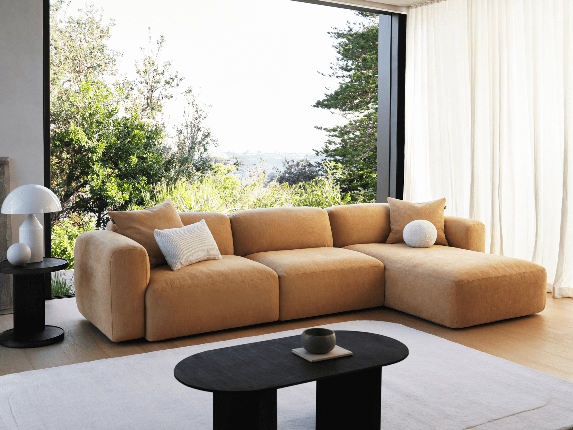 Luxurious modular sofa with plush camel-coloured upholstery, featuring deep seats and a chaise lounge. Set in a bright living space with large windows.