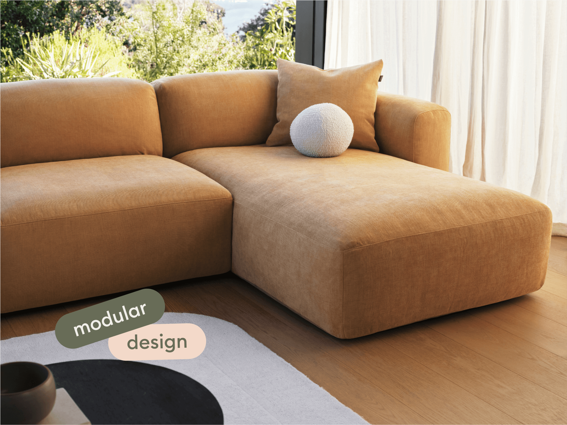 Modular sofa in plush tan fabric, featuring a chaise lounge. Set in a sunlit living room with floor-to-ceiling windows and light wood flooring.